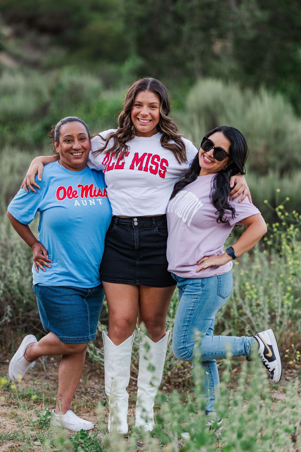 Three friends pose together outdoors wearing Ole Miss themed shirts and casual attire in a natural setting.