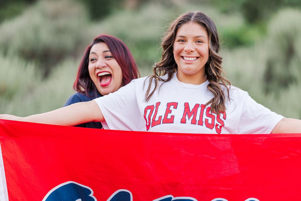 Two enthusiastic Ole Miss fans celebrate with a red flag while posing outdoors on a sunny day. graduation photos