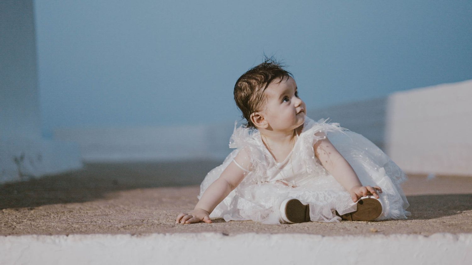 A small child in a delicate white dress sits on a light-colored carpet while looking to the side.