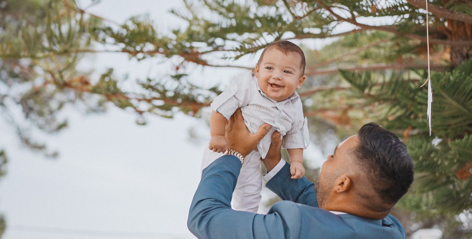 A delighted baby is lifted high in the air against a snowy winter landscape with evergreen trees.