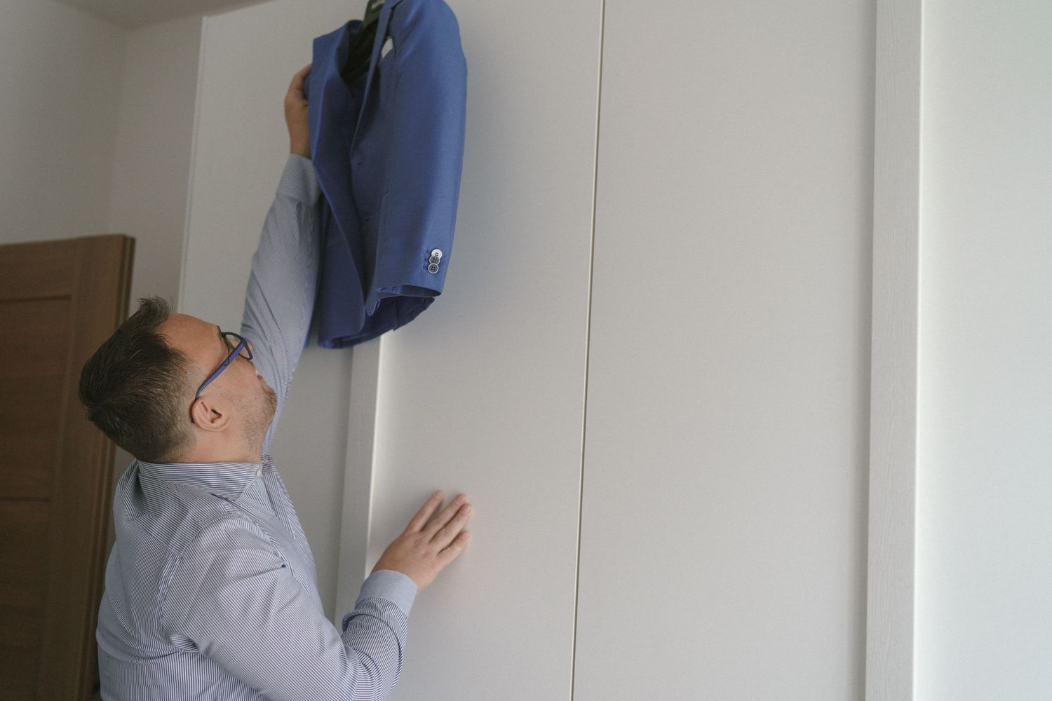 Person in glasses and gray shirt reaching up to inspect a blue backpack mounted on a white wall.