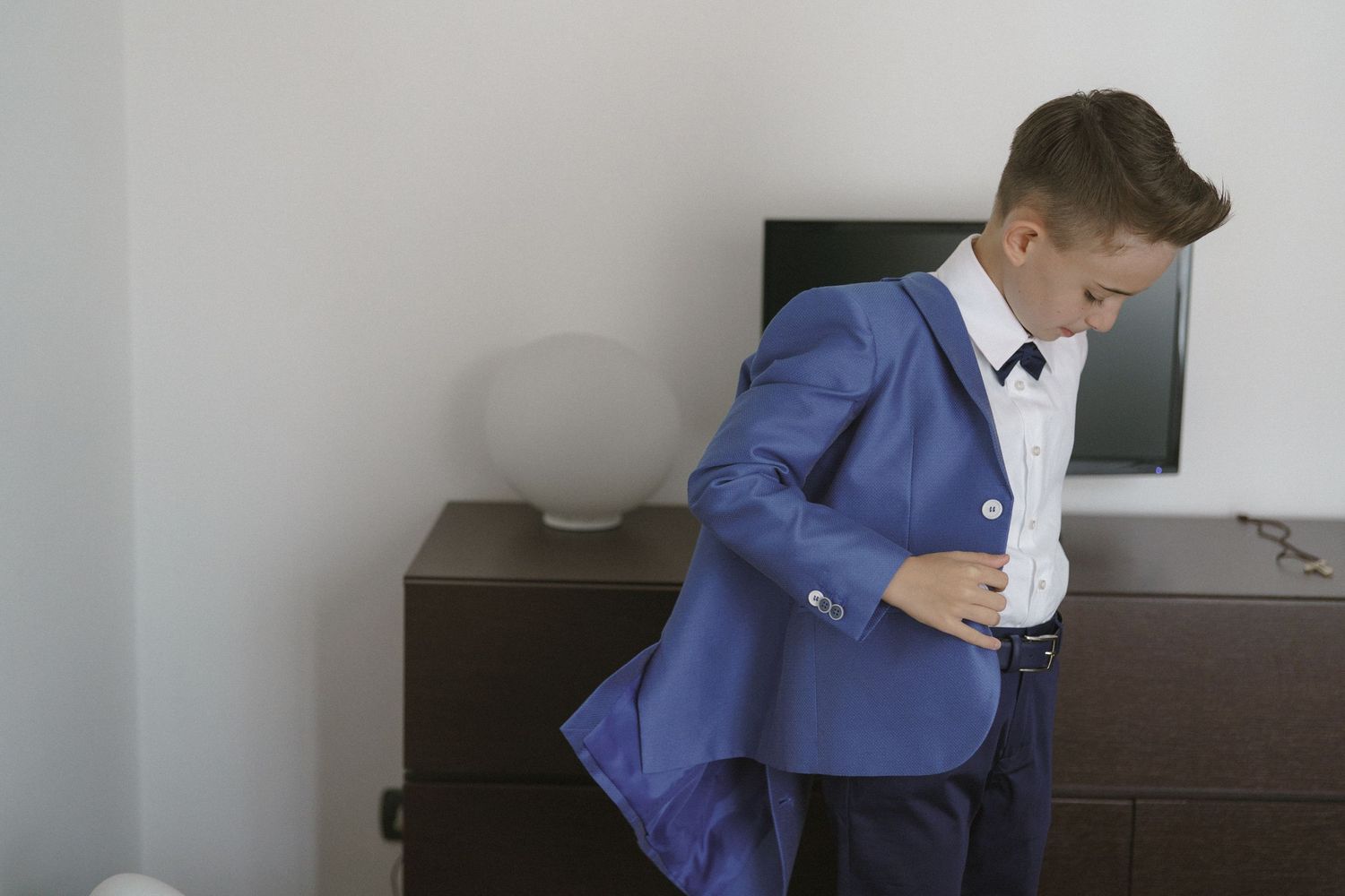 A person in a bright blue suit adjusts their jacket while standing next to a dark wooden dresser.