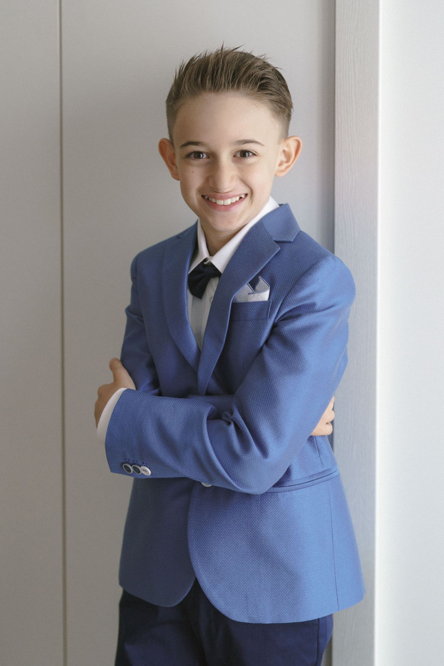 A young person in a blue blazer and black tie poses with crossed arms against a light wall.
