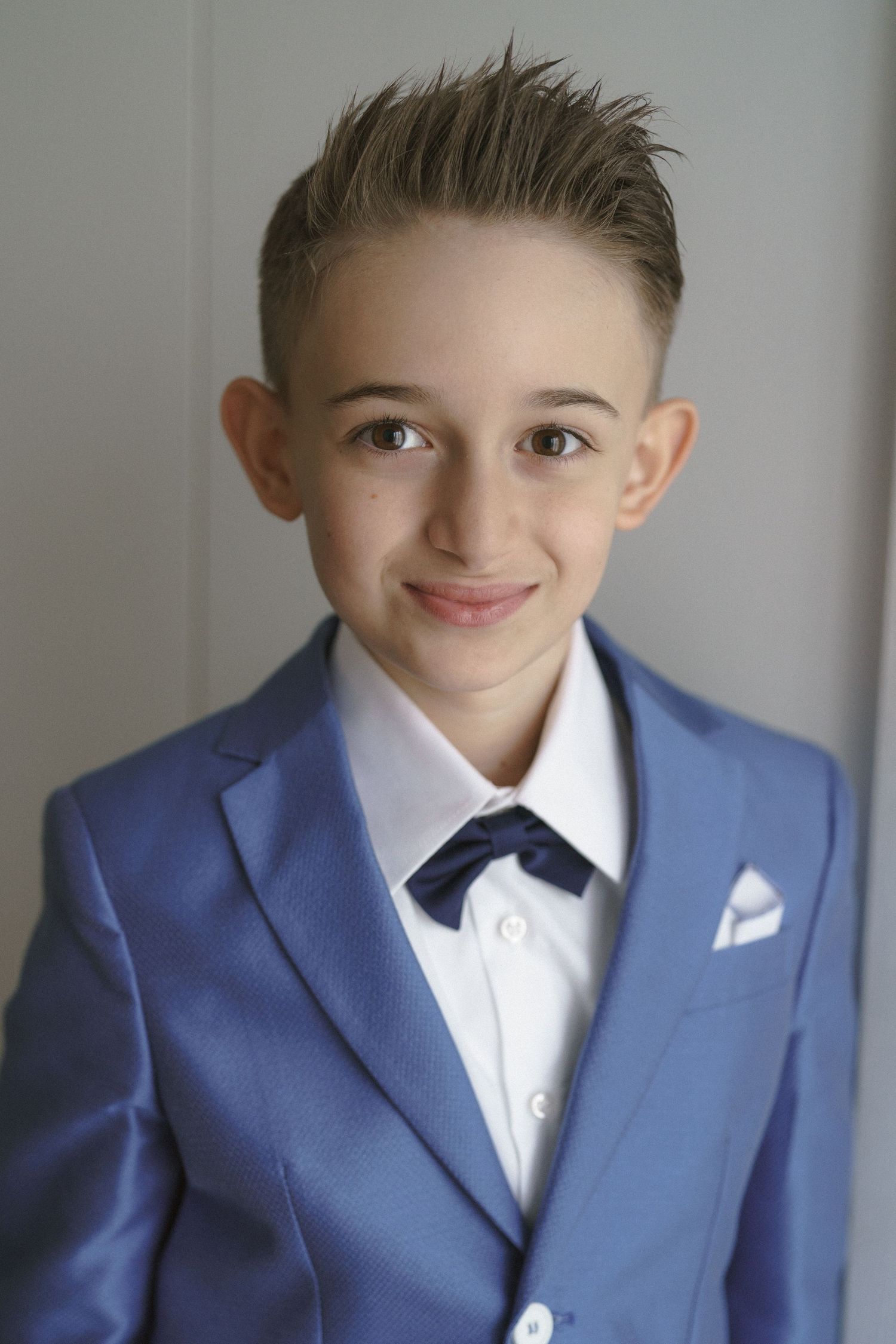 Young child wearing a crisp blue suit jacket with white dress shirt and navy tie in a formal portrait setting.