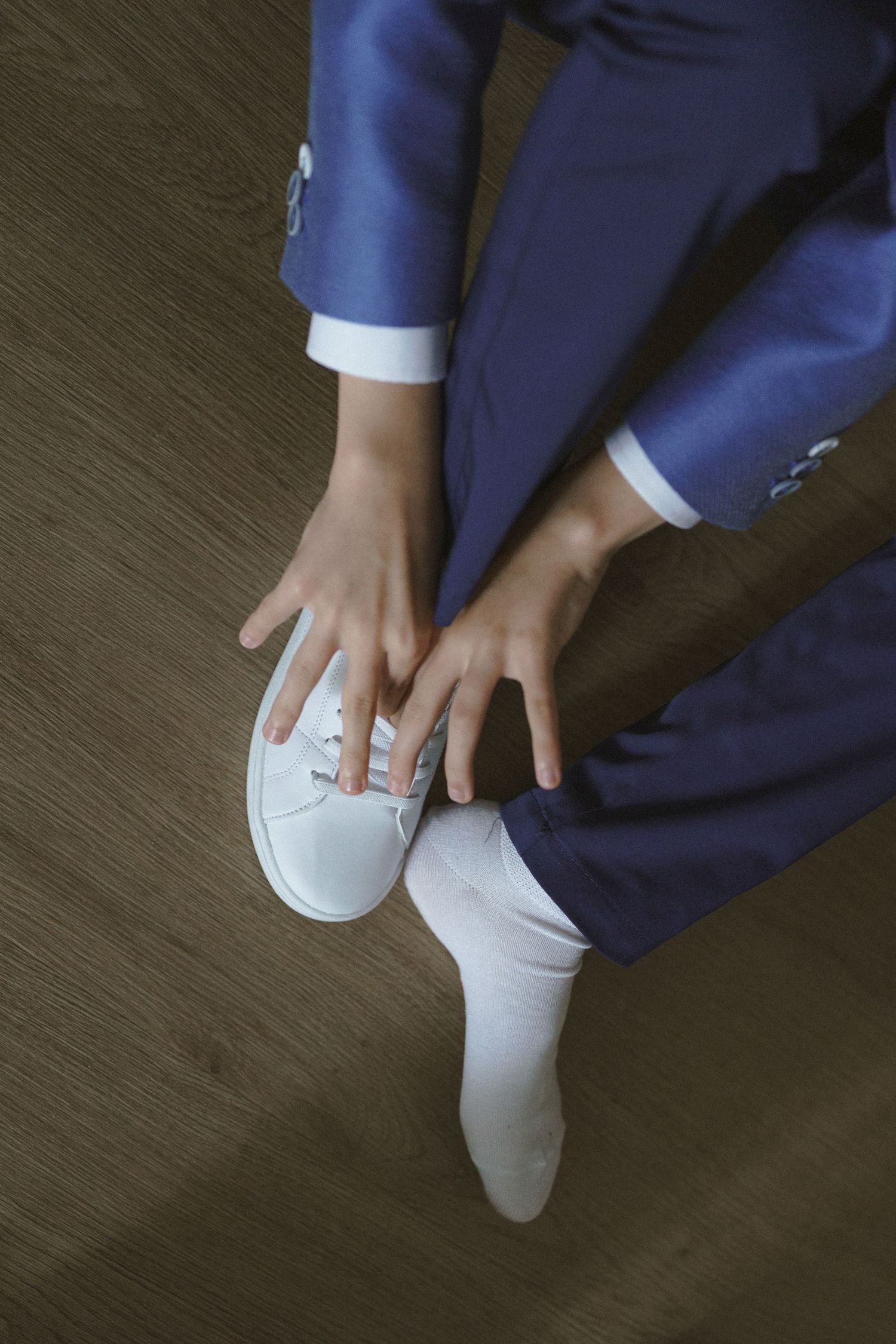 Hands adjusting white dress shoes with navy blue suit and white socks on wooden floor, viewed from above.