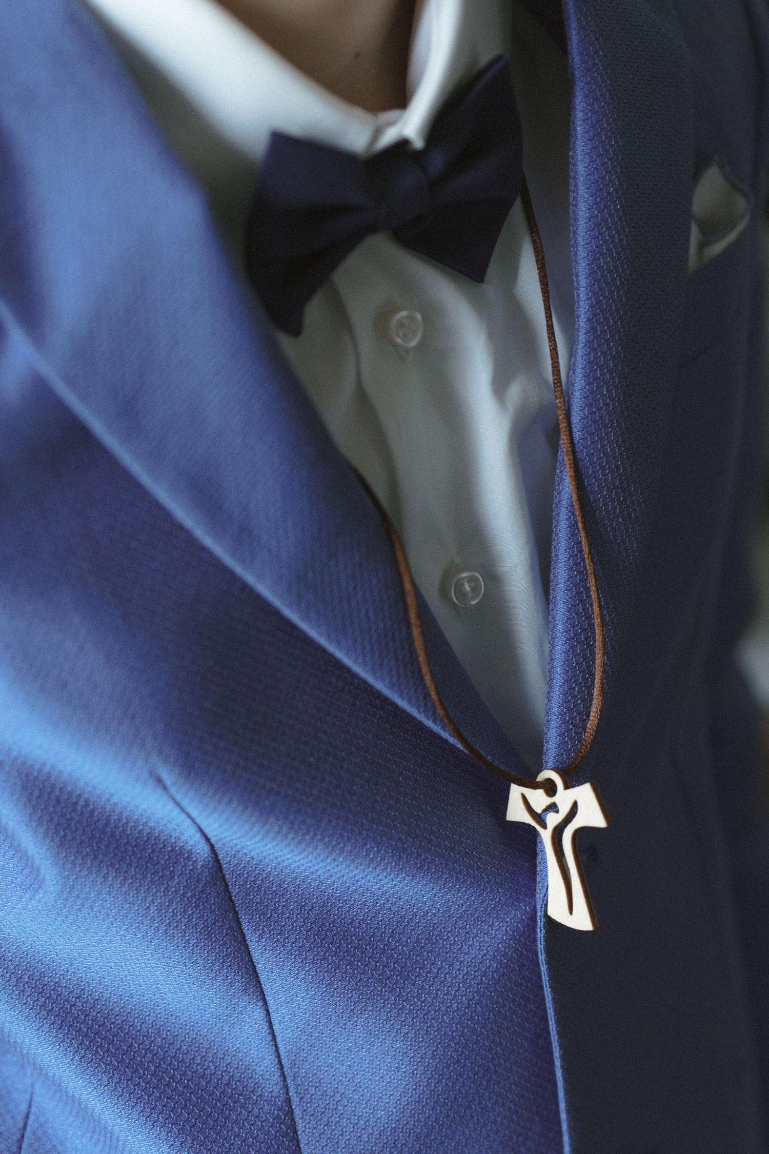 Close-up of a navy blue suit jacket with black bow tie and silver cross necklace pendant against white dress shirt.