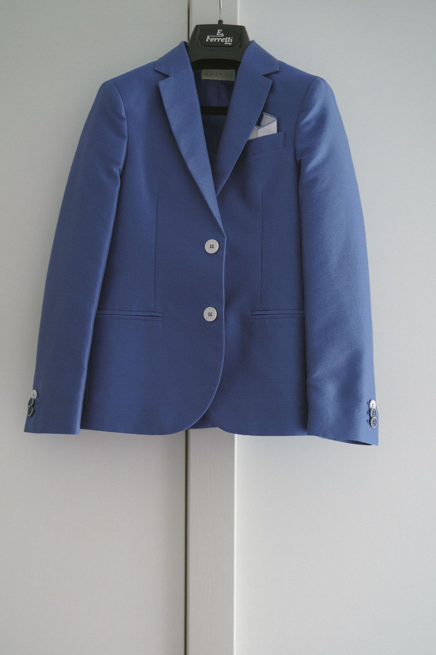 A blue tailored suit jacket with white buttons and a white pocket square hangs on a hanger against a light wall.