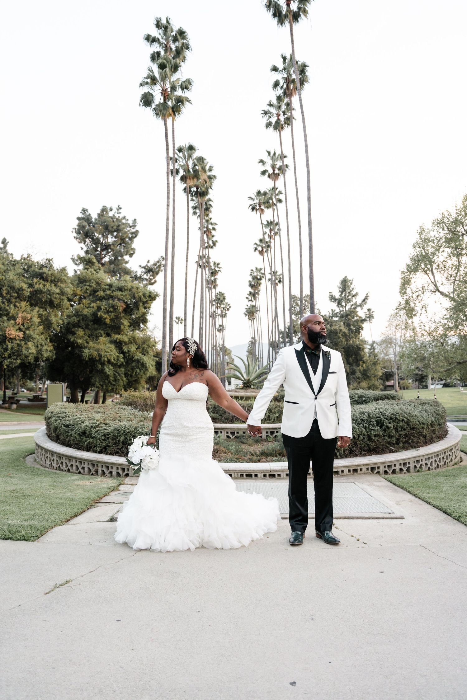 Newlyweds hold hands at golden hour under towering palm trees in a scenic garden setting.