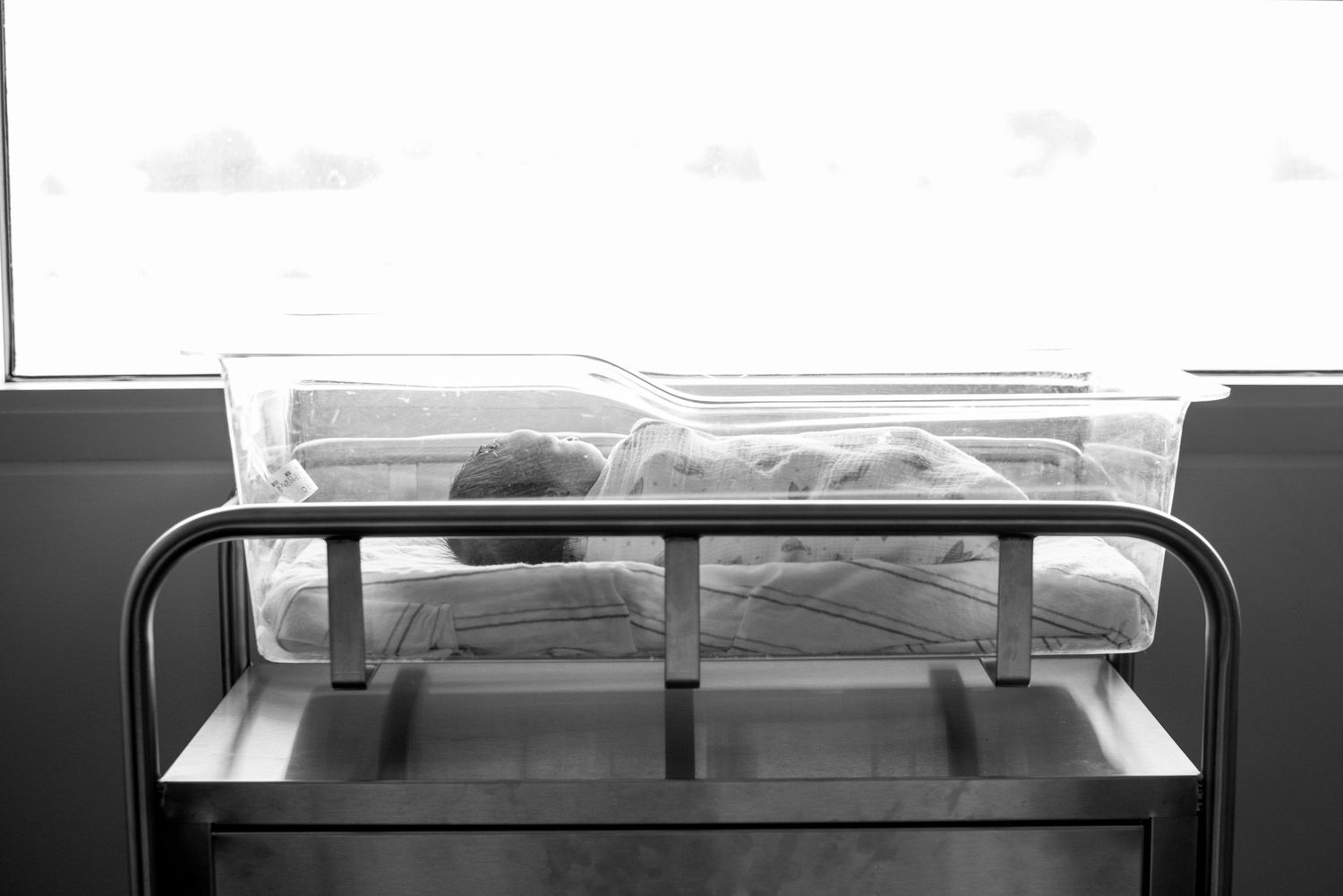 A black and white photograph of a newborn in a clear bassinet near the hospital window.  Fresno Fresh 48 Hospital Photographer