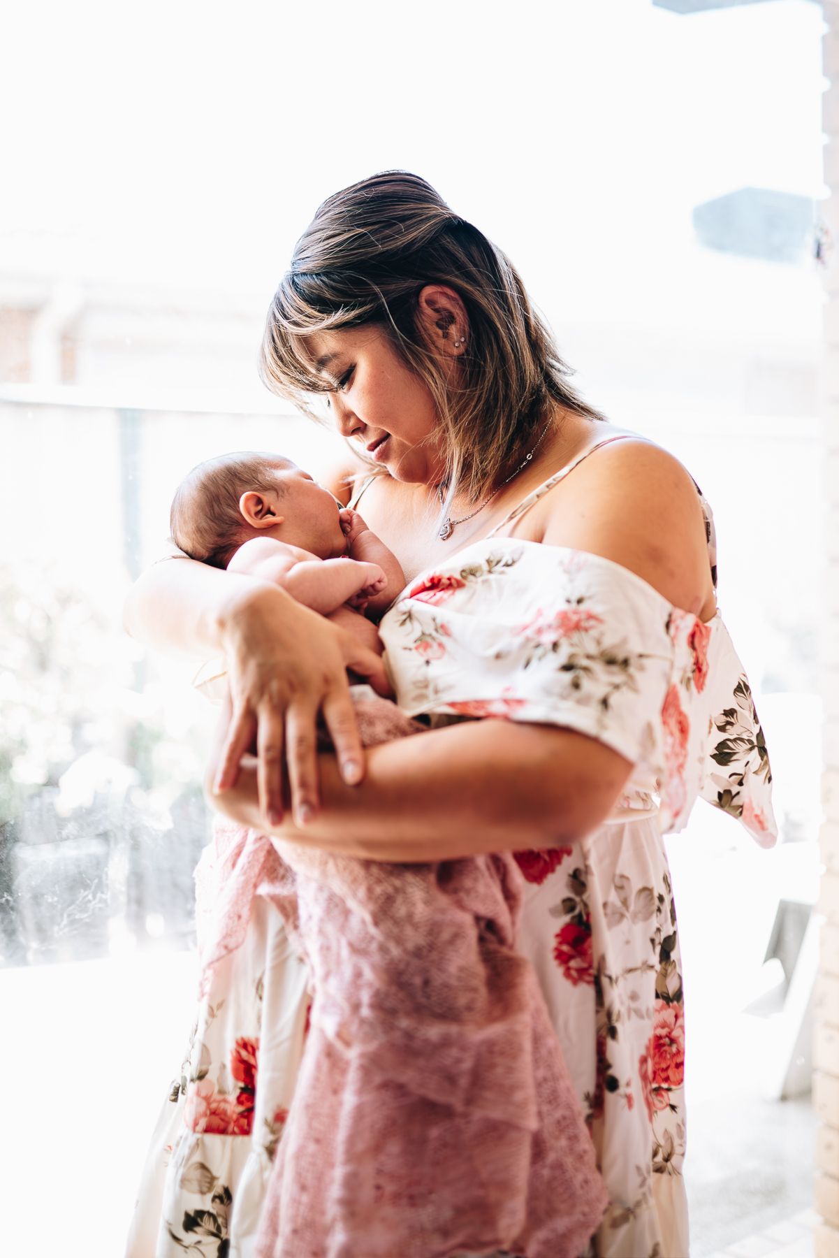 A tender moment between mother and newborn in a floral dress by sunlit window taken by Melbourne newborn maternity photographer