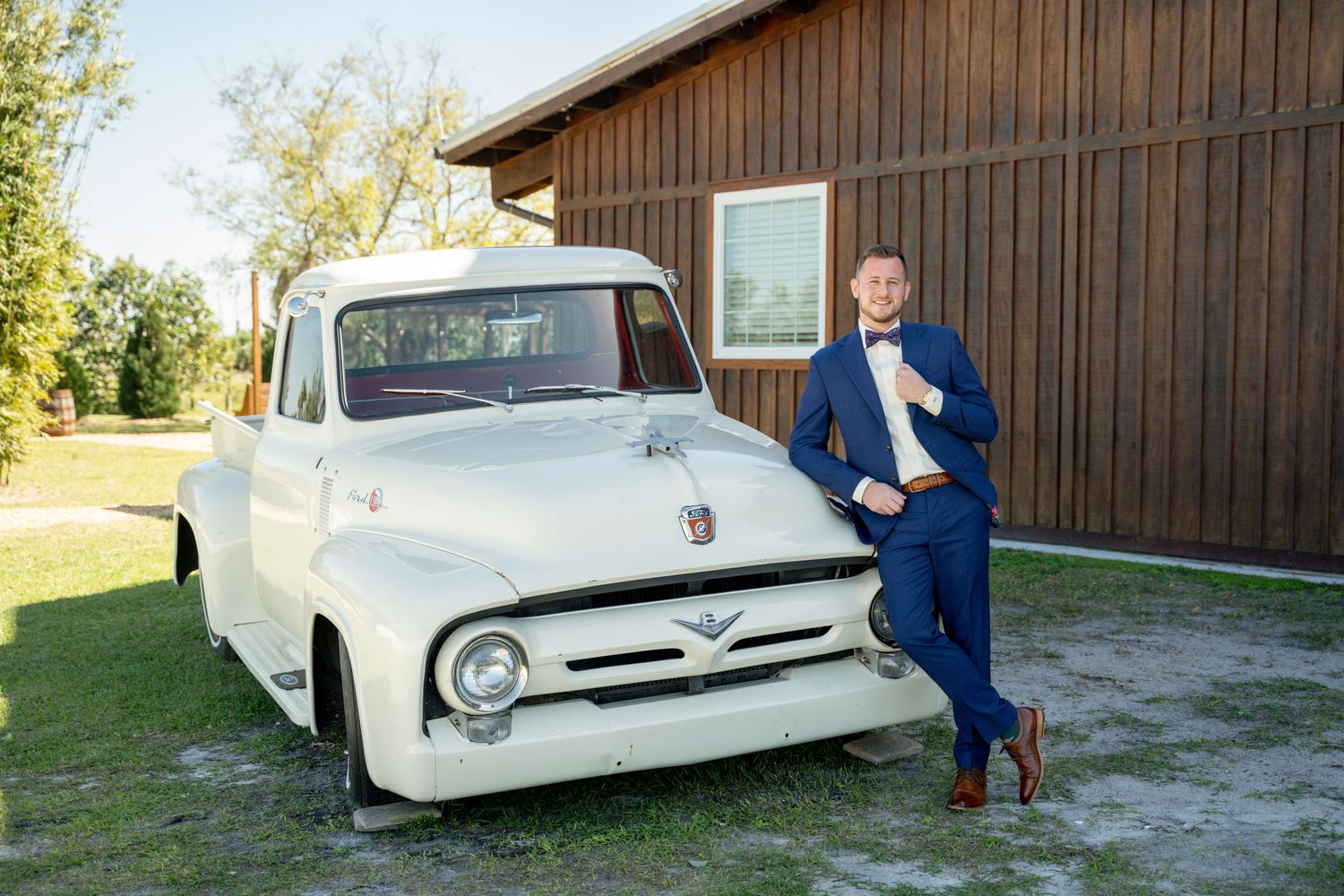 Classic white Ford pickup truck parked beside wooden barn in rural setting.