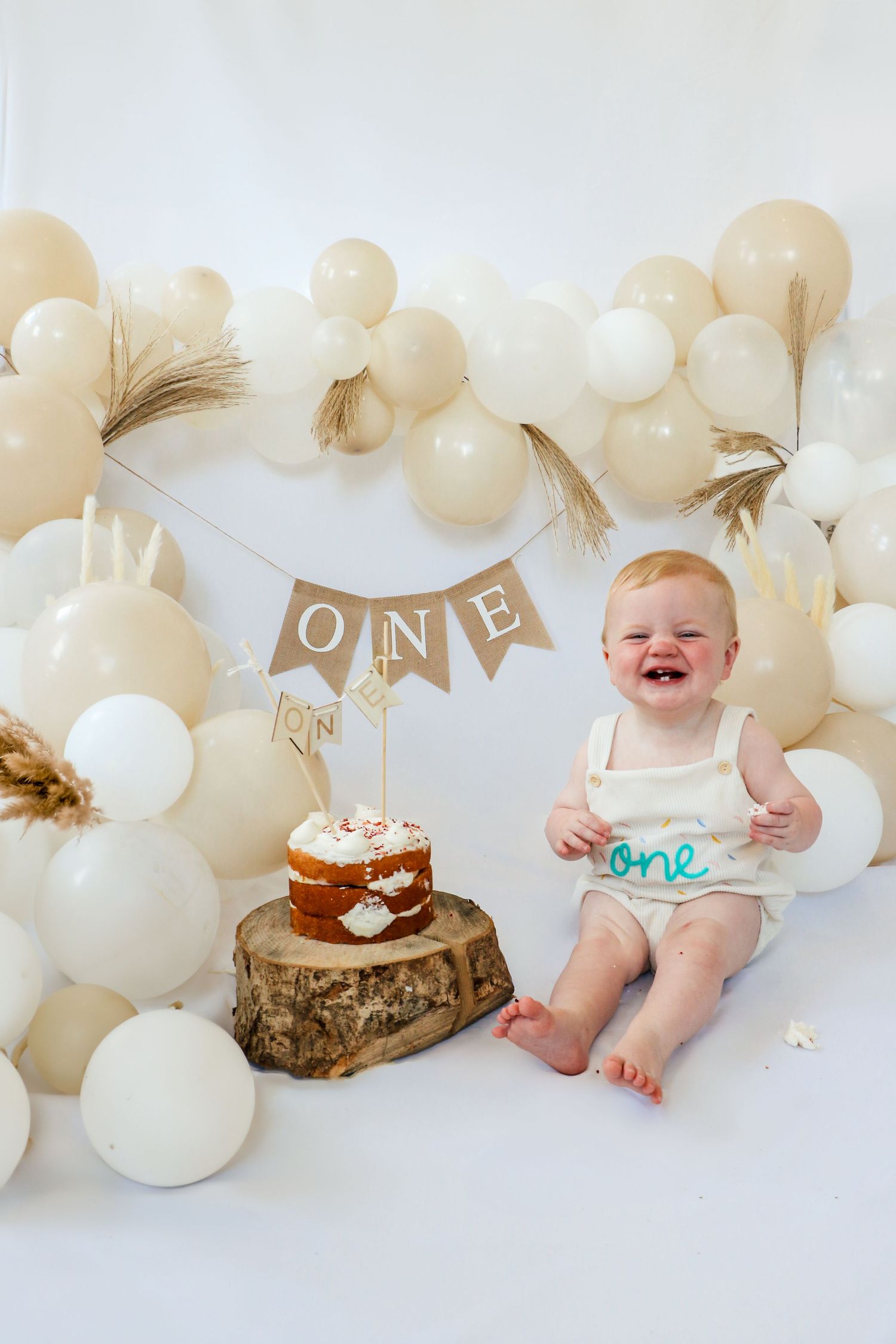 Baby wearing crown and white dress poses with birthday cake surrounded by balloons. Coventry Cake Smash Photographer. First Birthday.