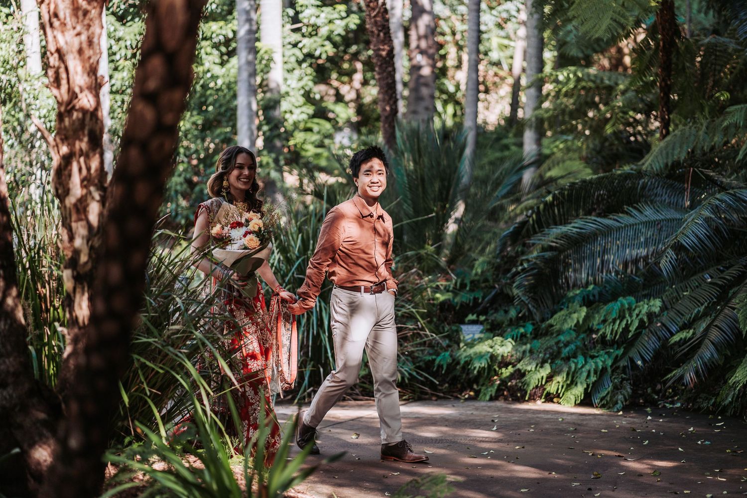 People walking on a scenic forest trail surrounded by lush ferns and towering redwood trees in dappled sunlight.