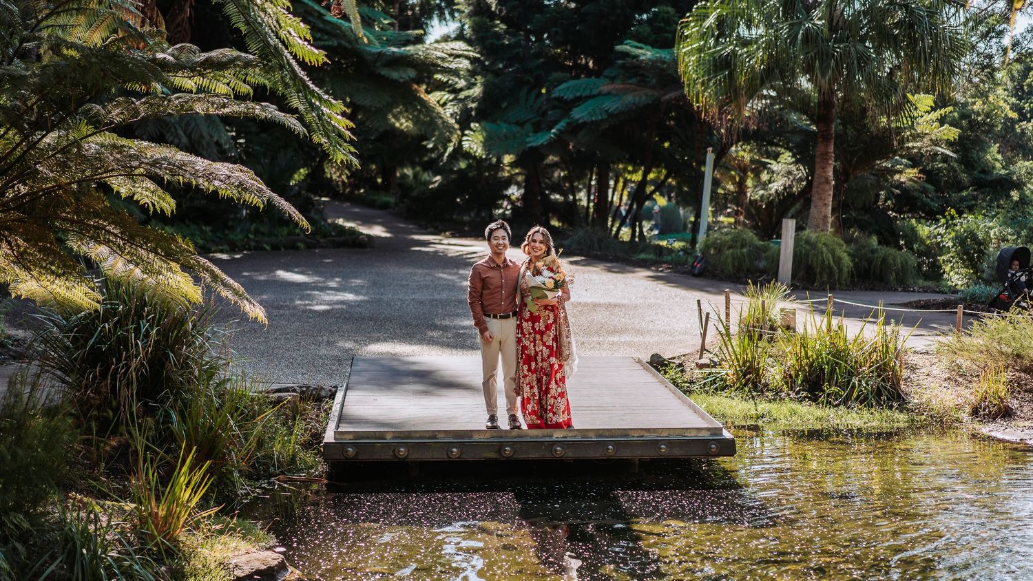 Two people in floral attire stand together on a wooden dock by a tranquil pond surrounded by lush gardens and trees.