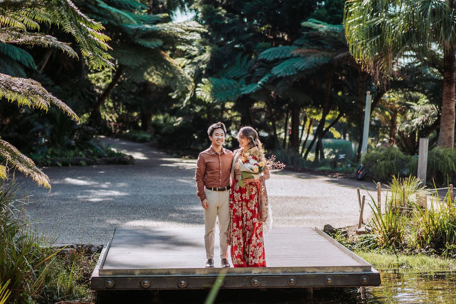 A couple walks along a scenic wooden pathway surrounded by lush tropical plants and palm trees in a garden setting.