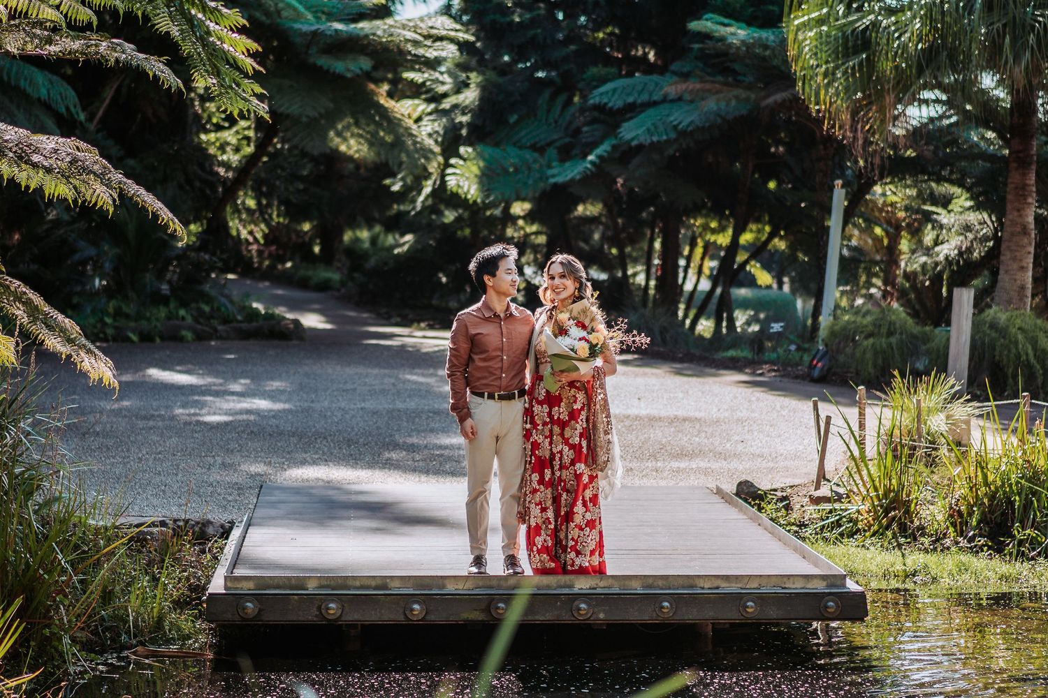 A couple stands together on a wooden bridge surrounded by lush tropical gardens and palm trees on a sunny day.