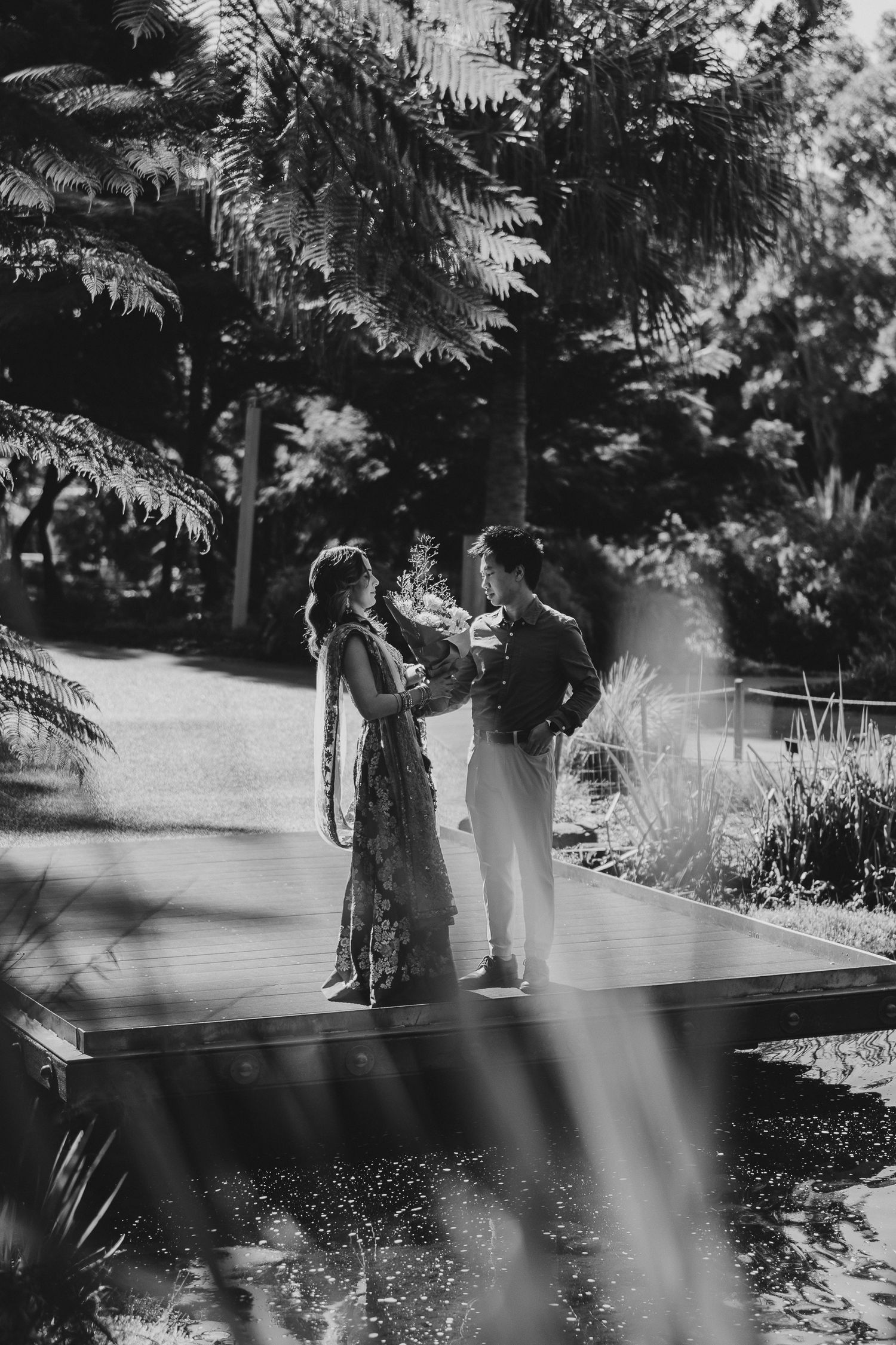 A romantic black and white photo of a couple standing in a sunlit garden with tropical foliage and light beams.