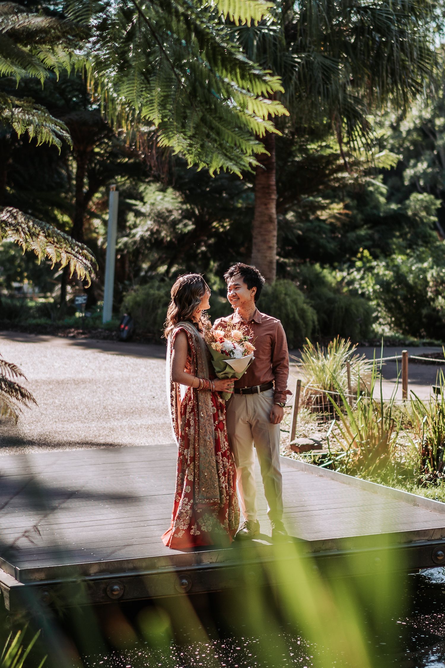 Romantic couple stands together in a lush garden setting with palm trees and greenery on a sunny afternoon.