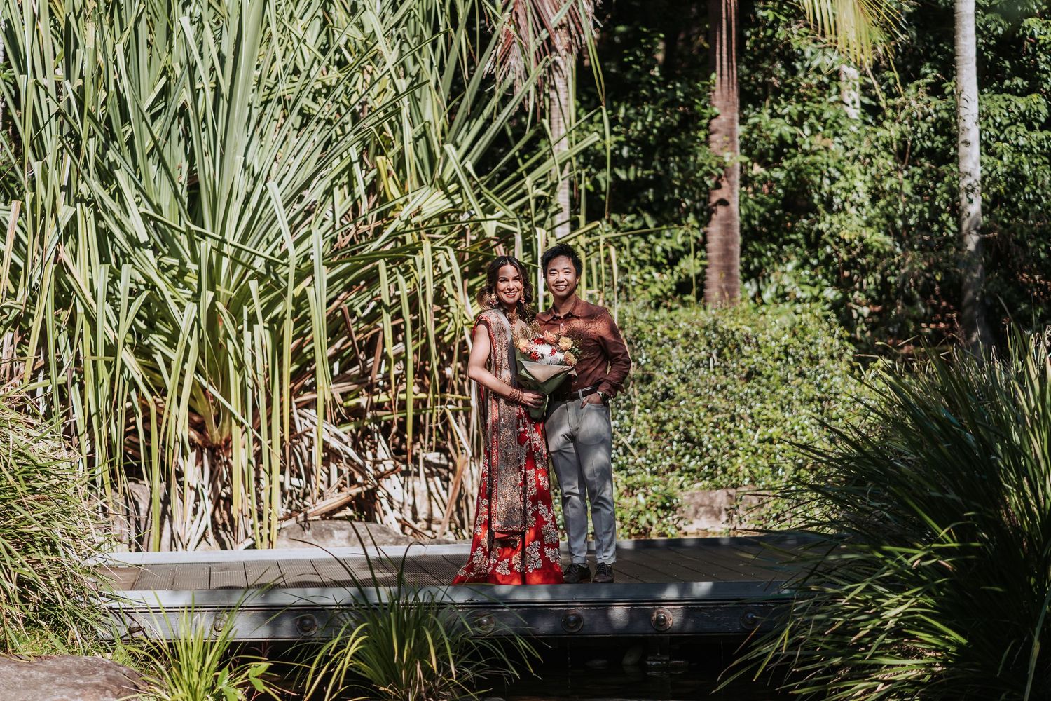 A couple poses together on a wooden deck surrounded by lush tropical plants and tall grass in a garden setting.