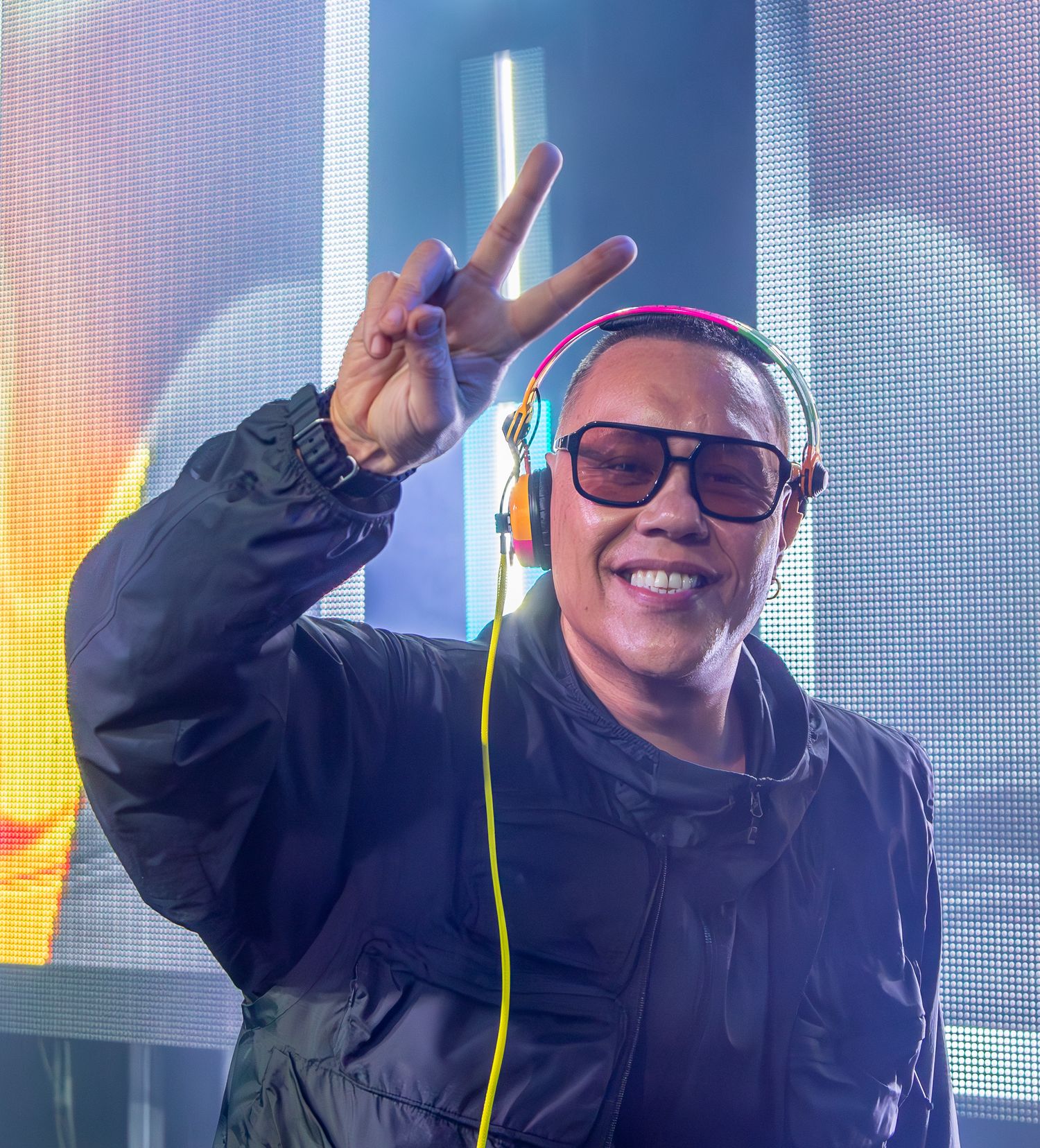 DJ in black jacket and sunglasses smiles while giving peace sign during colorful performance on stage.