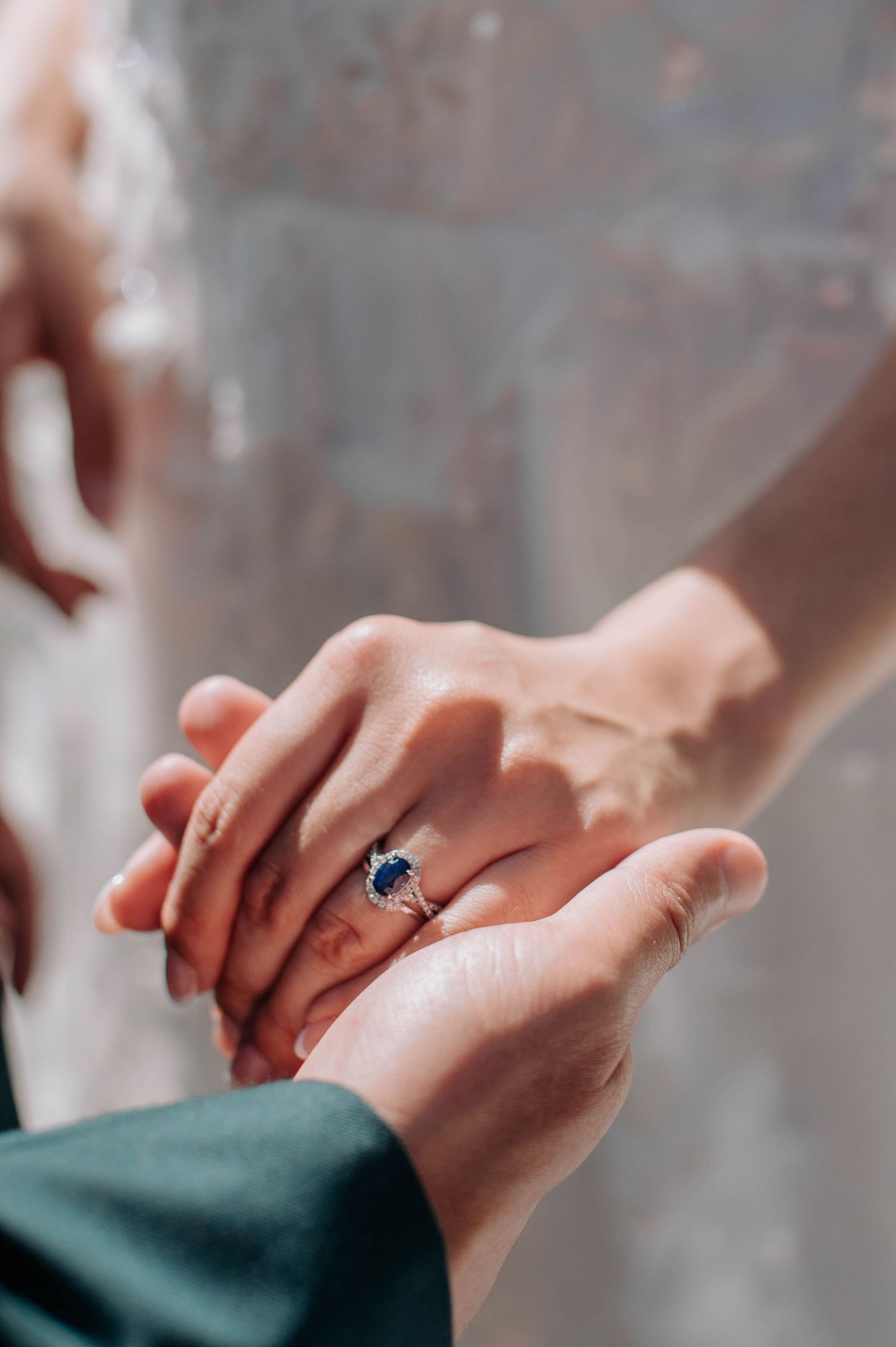 Close-up shot of hands showing off a stunning sapphire engagement ring against a blurred gray background.