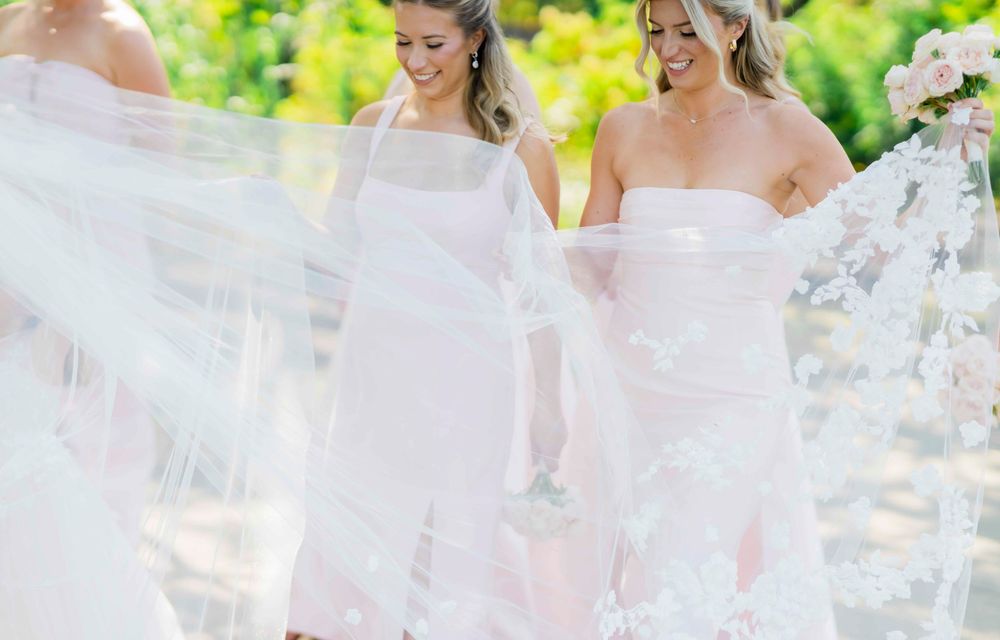 Two people wearing elegant white off-shoulder dresses walk gracefully through an outdoor garden setting.