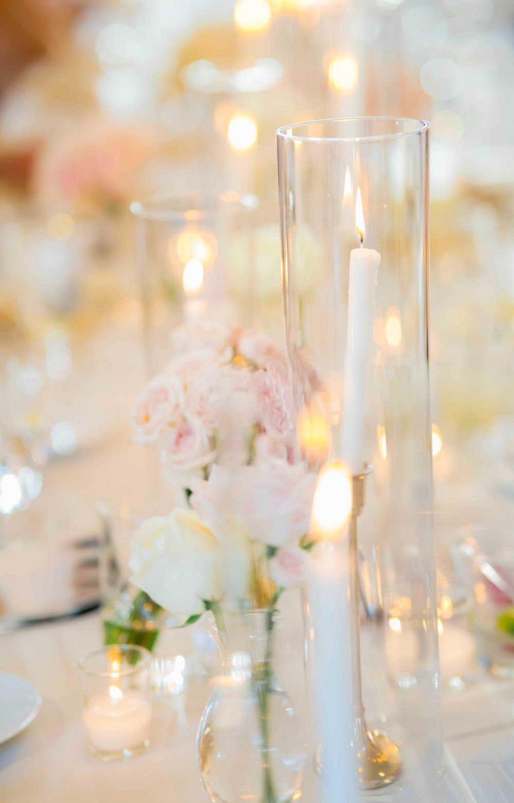 Elegant wedding centerpiece with tall glass candleholder and soft pink roses on white tablecloth with glowing bokeh lights.