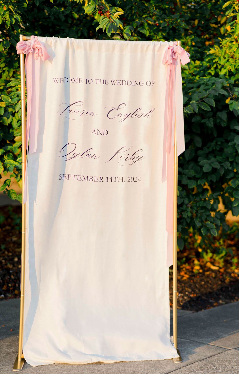 Elegant white wedding welcome sign with blush pink ribbons on gold stands against a green garden backdrop.