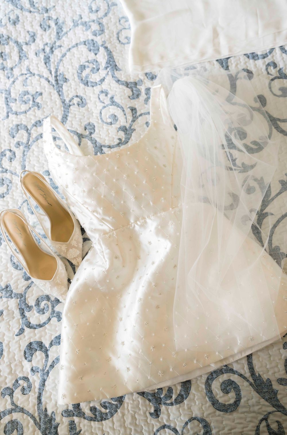 Elegant white wedding dress with delicate pearl details laid out on a blue and white patterned background with matching shoes.