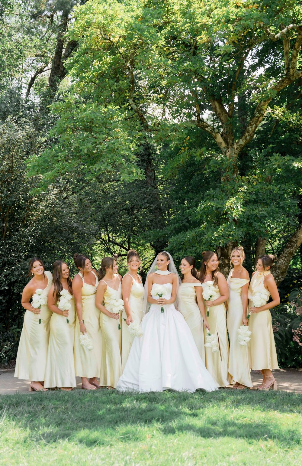 Wedding party in yellow bridesmaid dresses and white bridal gown pose together outdoors in a sunny garden setting.