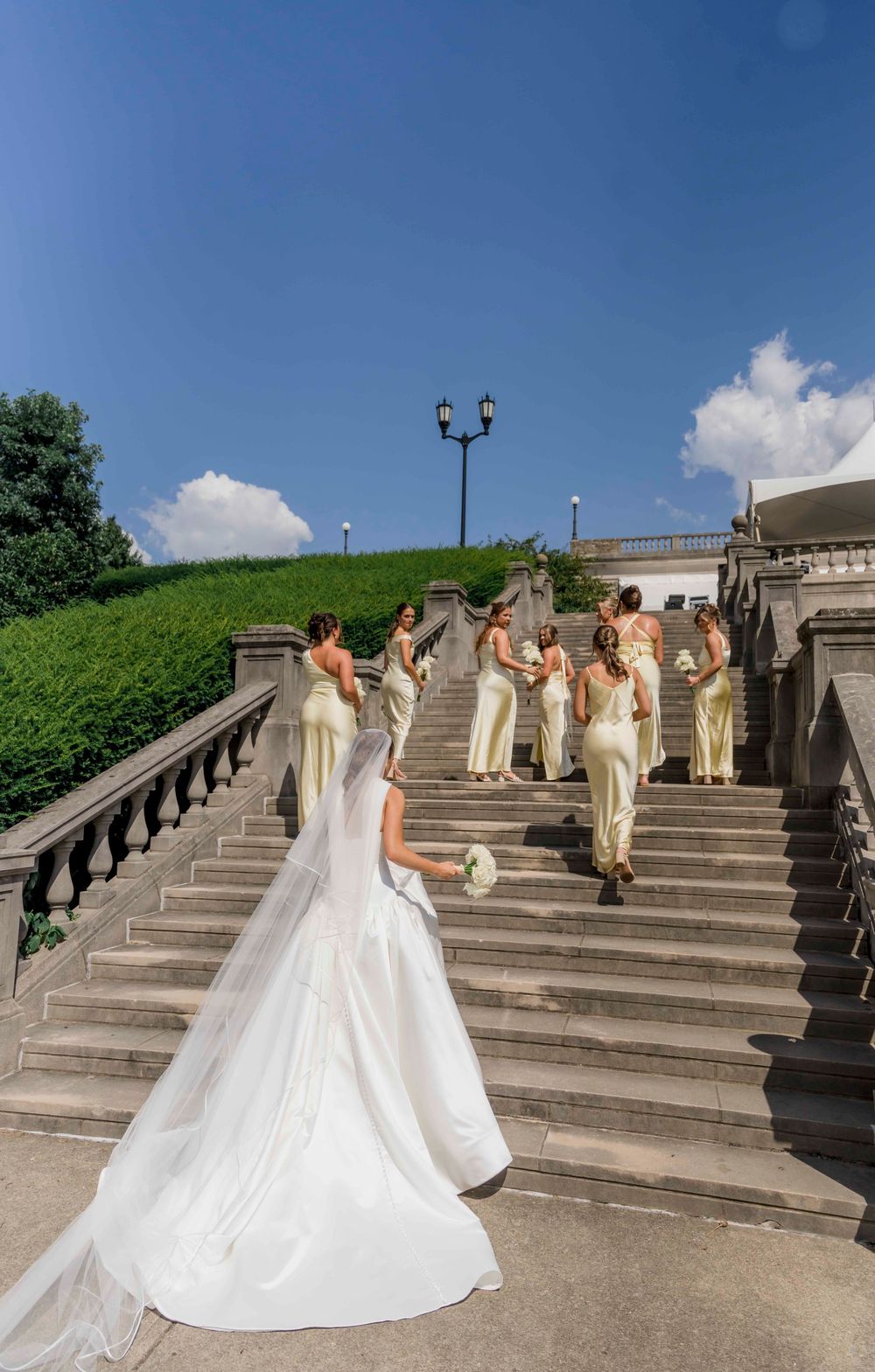 Wedding party members in yellow dresses follow the bride up grand stone stairs on a sunny summer day.