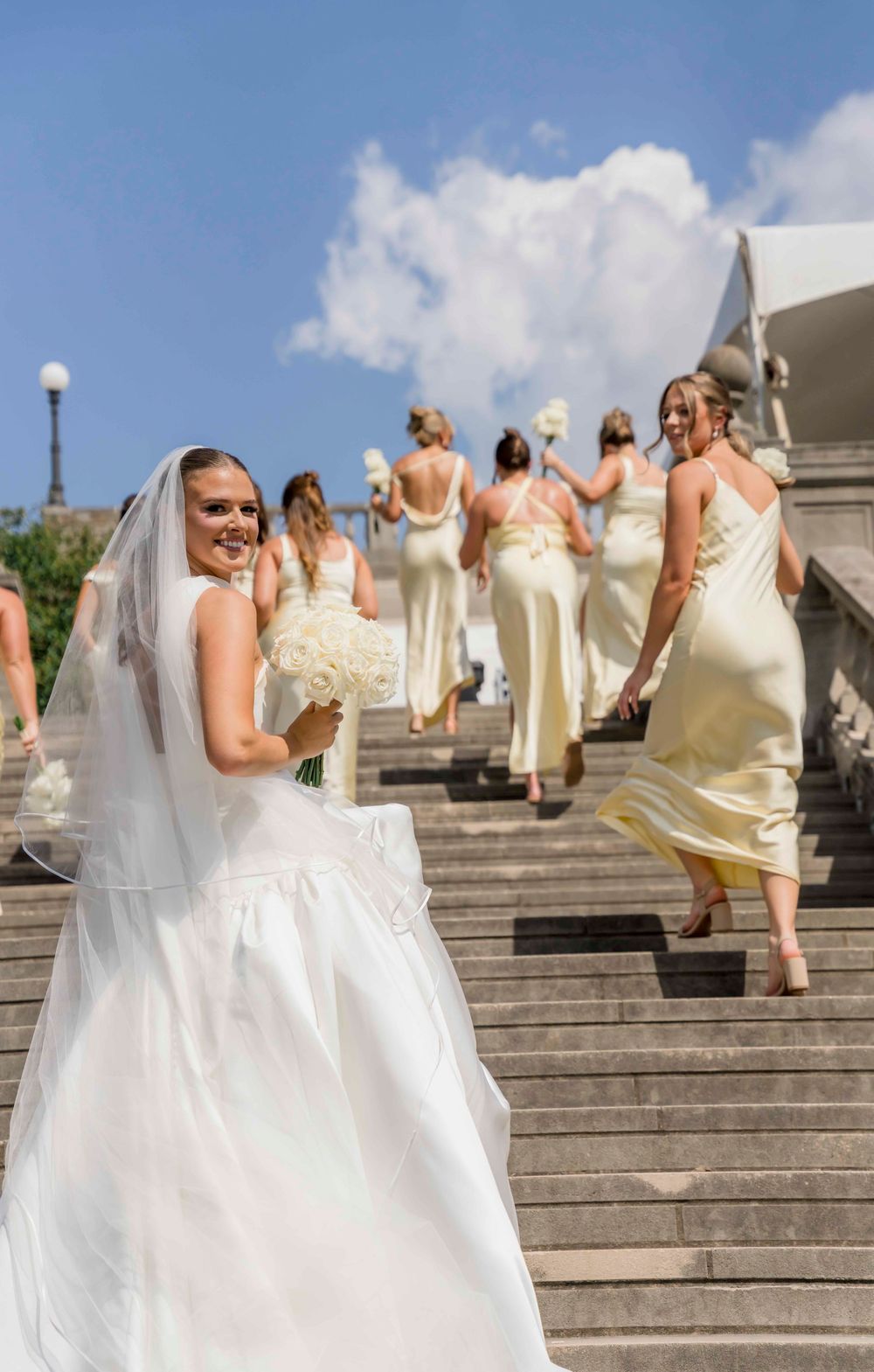 Wedding party in champagne-colored dresses walking up stone steps while bride holds white bouquet in foreground.