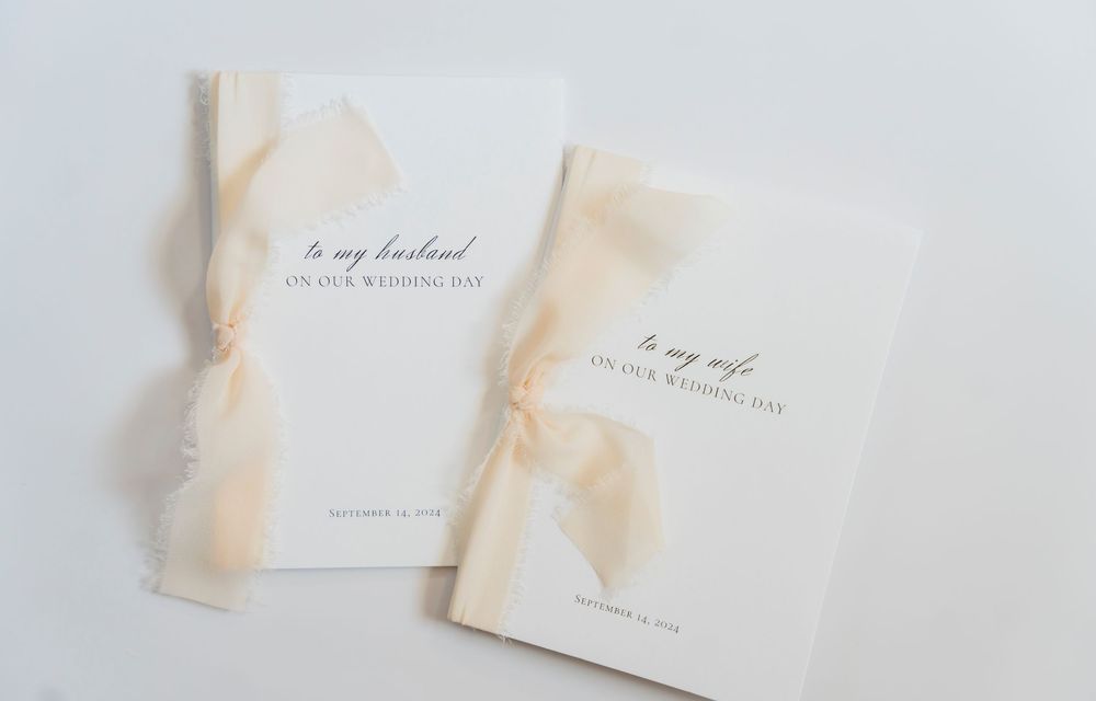 Two elegant wedding vow books with cream-colored covers and delicate peach ribbons lay side by side on white surface.