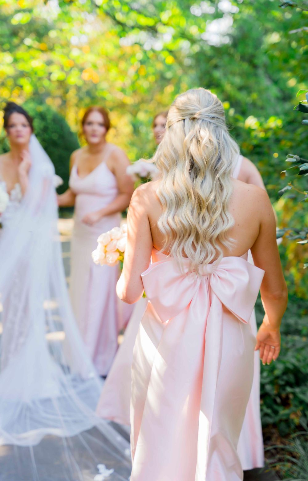 Bridesmaids in flowing pink dresses gathered outdoors with delicate bow details and elegant hairstyles.