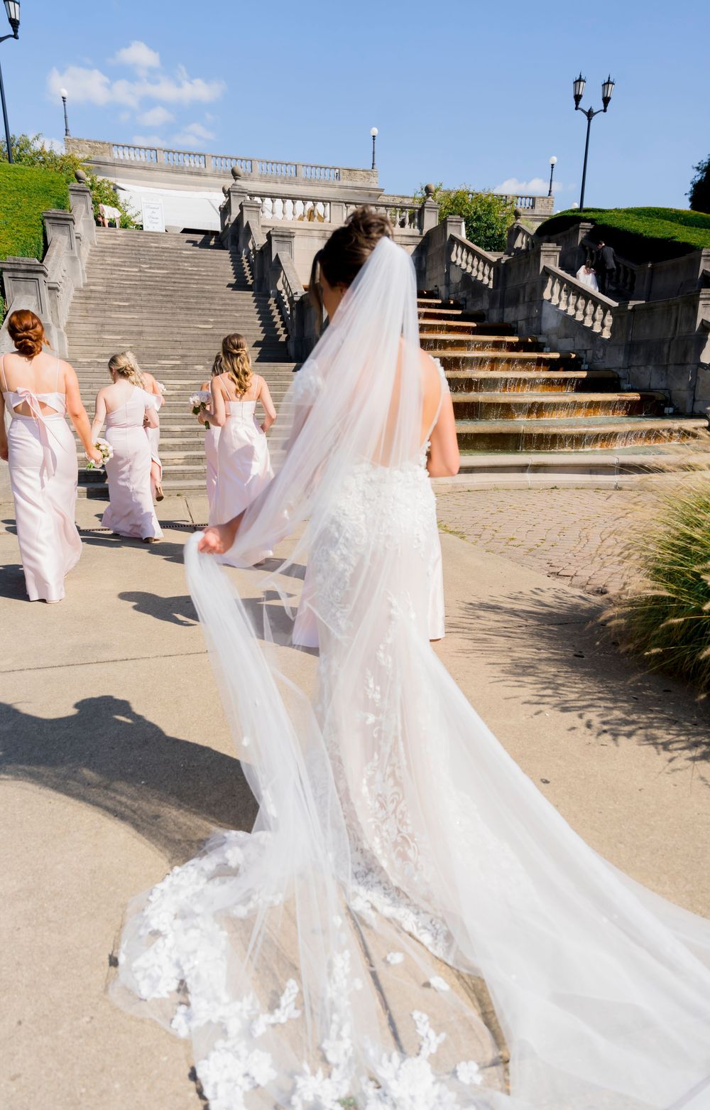 Bride in flowing white lace gown with long veil walks up grand outdoor stone staircase with bridesmaids.