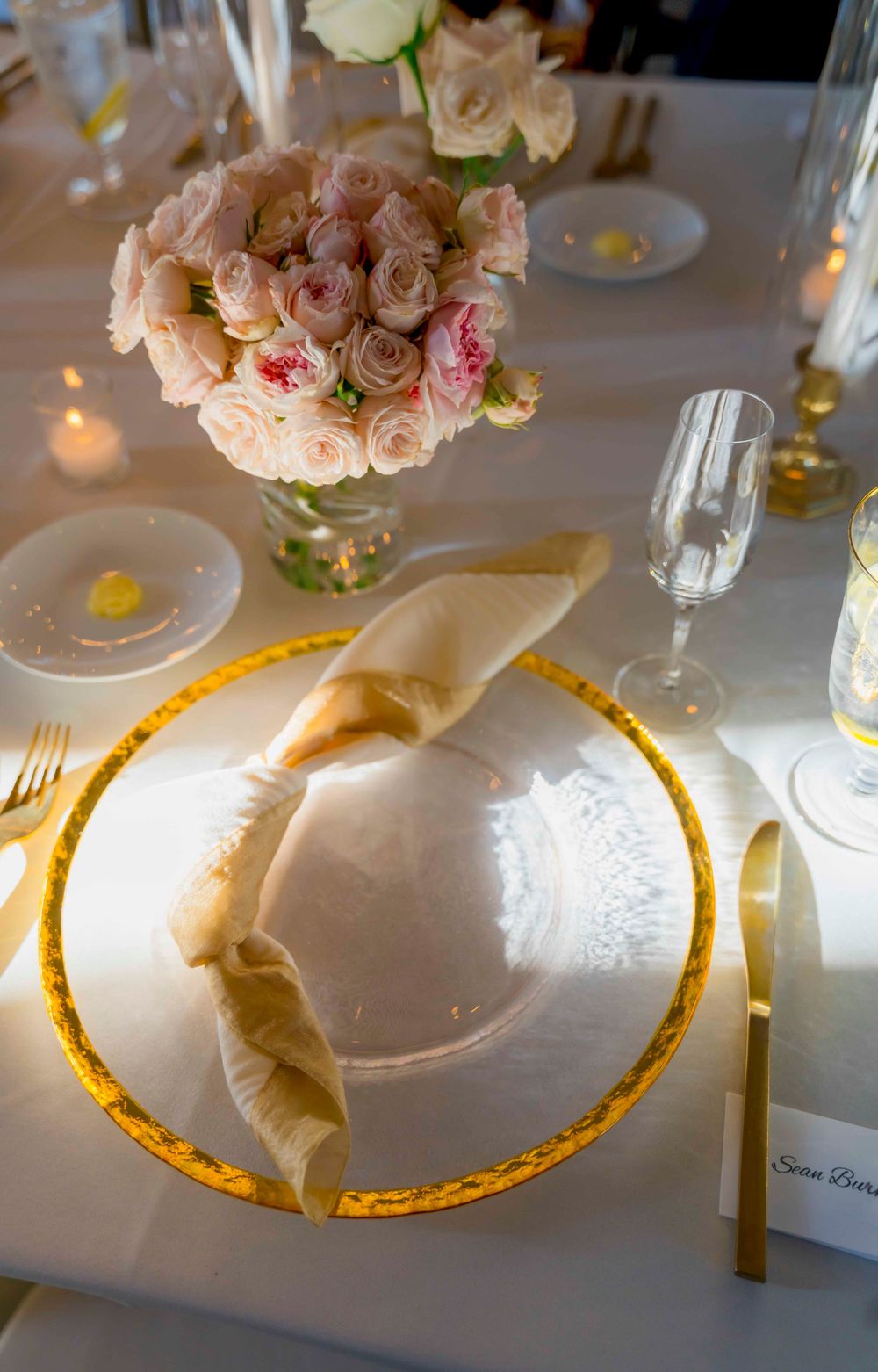 Elegant table setting with gold-rimmed plates, white napkins, and soft pink rose centerpiece arrangements in ambient lighting.