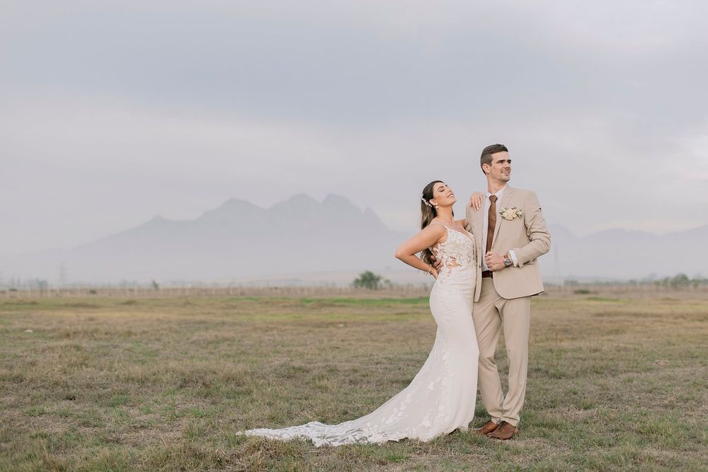 Bride and groom stand elegantly in an open field with a dramatic mountain backdrop on their wedding day.