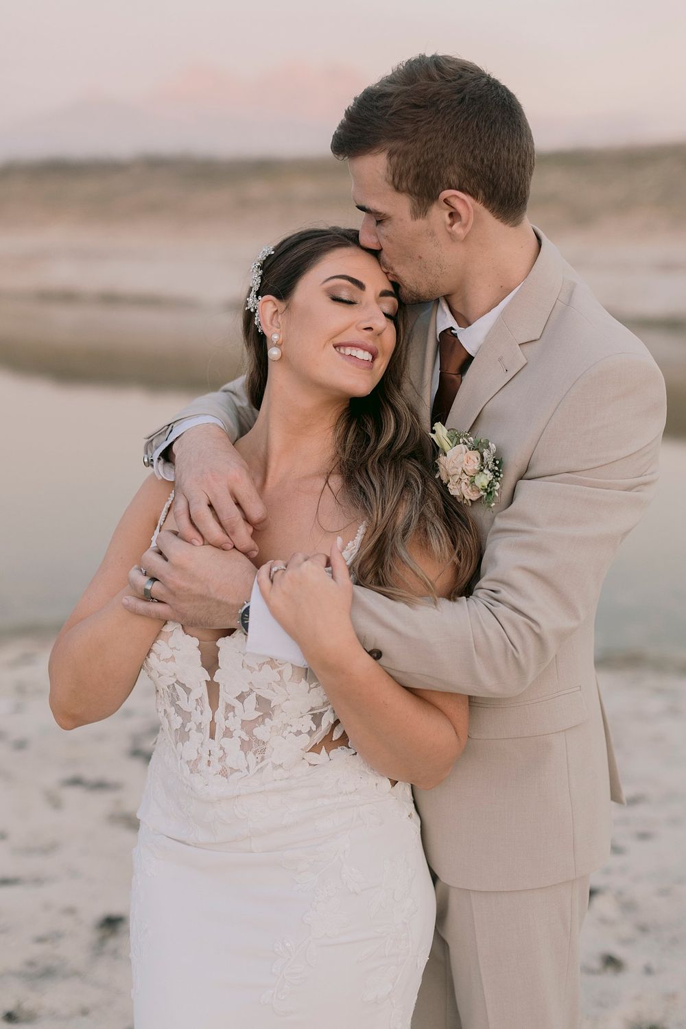 Groom in beige suit kisses bride in lace wedding dress during golden hour outdoor portrait session.