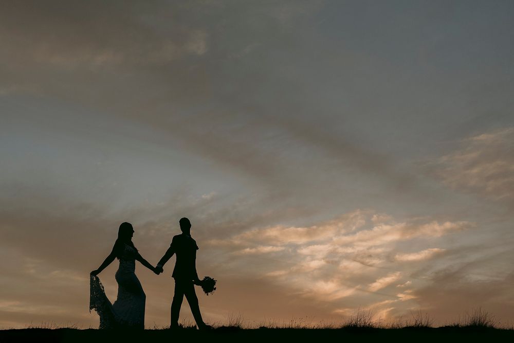 Silhouette of bride and groom holding hands against a dramatic sunset sky with wispy clouds.