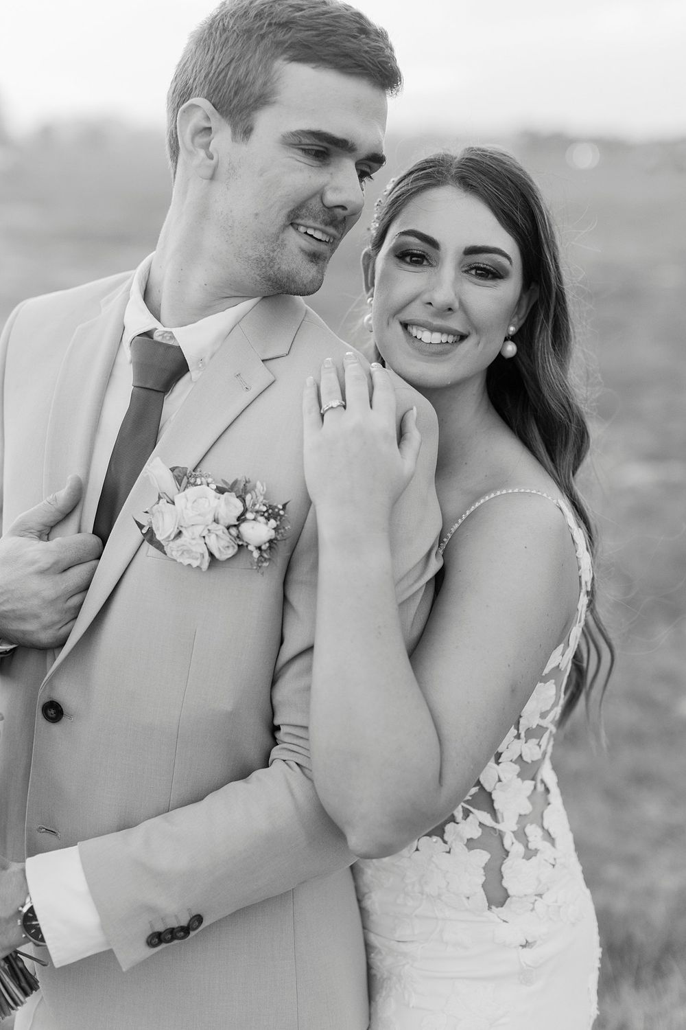 Black and white portrait of smiling bride in lace dress leaning against groom in beige suit.
