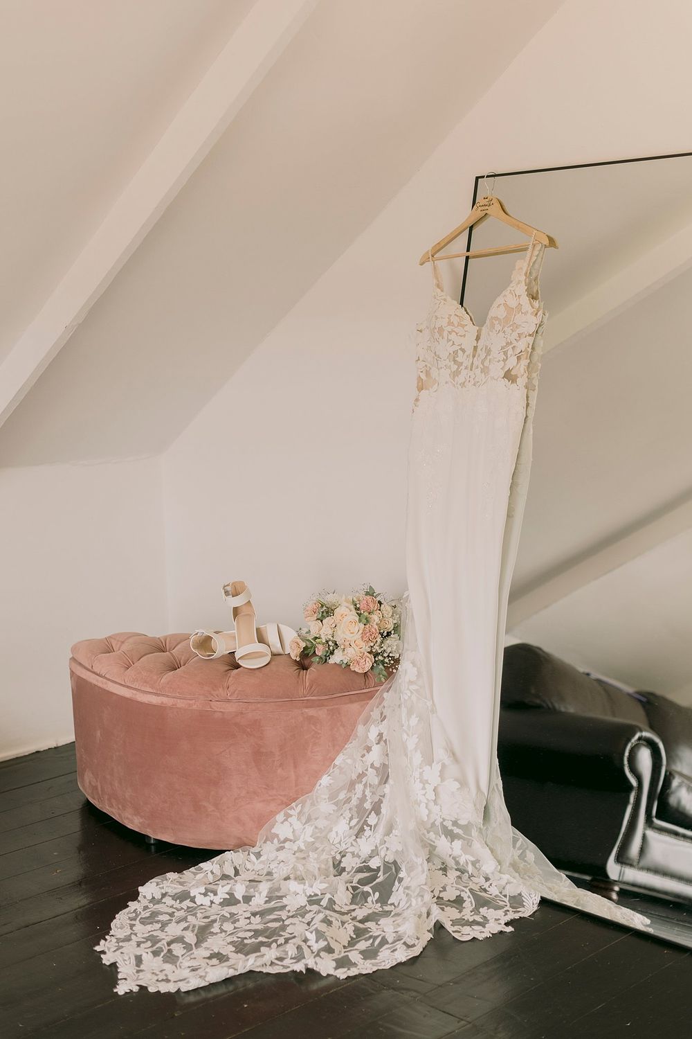 Elegant lace wedding gown with long train hanging on a mirror beside a pink velvet ottoman and bridal bouquet.