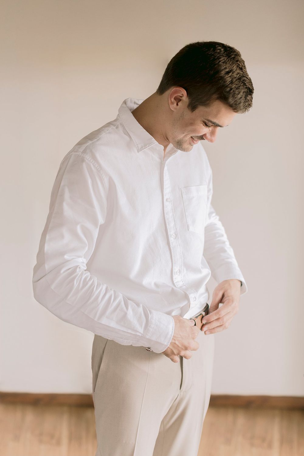Smiling groom in white dress shirt and khaki pants adjusts his belt while getting ready for his wedding day.