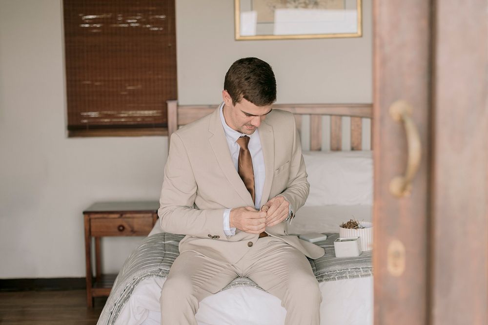Groom in tan suit sitting on bed buttoning jacket during wedding morning prep.