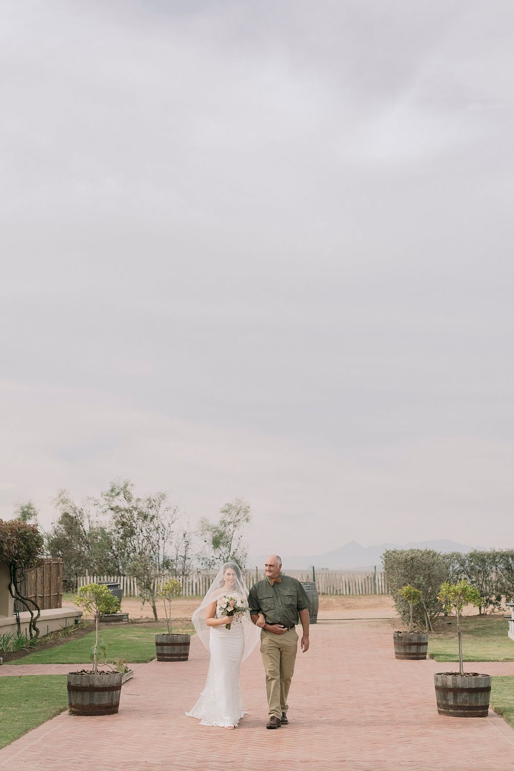 Bride in white gown walks down outdoor brick aisle with escort under cloudy sky at vineyard venue.