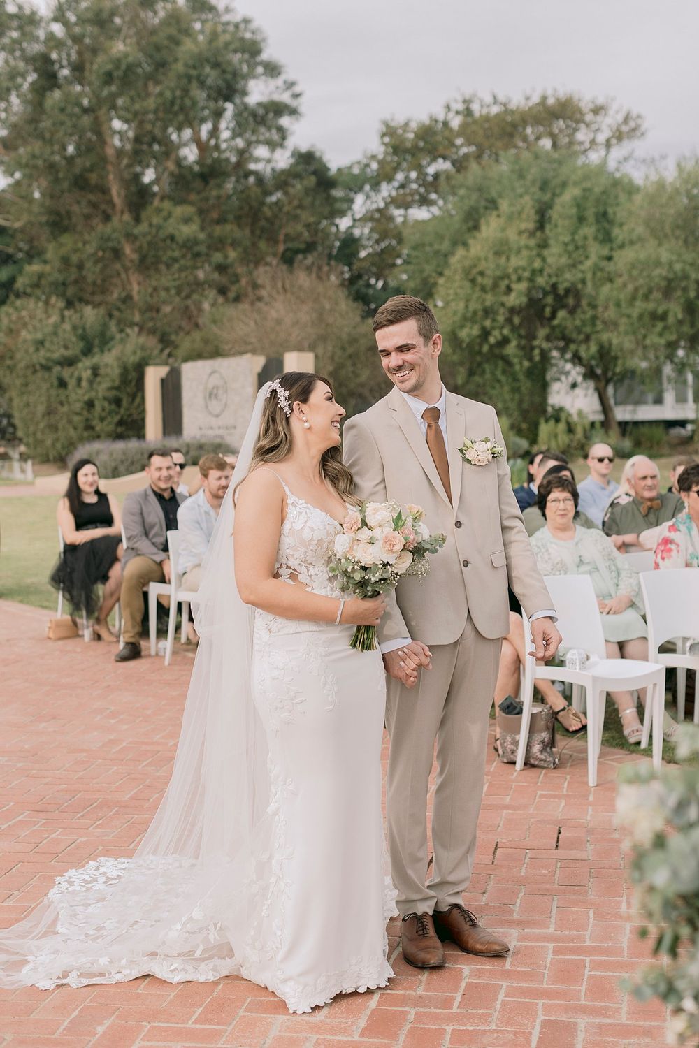 Newlyweds stand hand in hand smiling at outdoor ceremony with seated wedding guests behind them.
