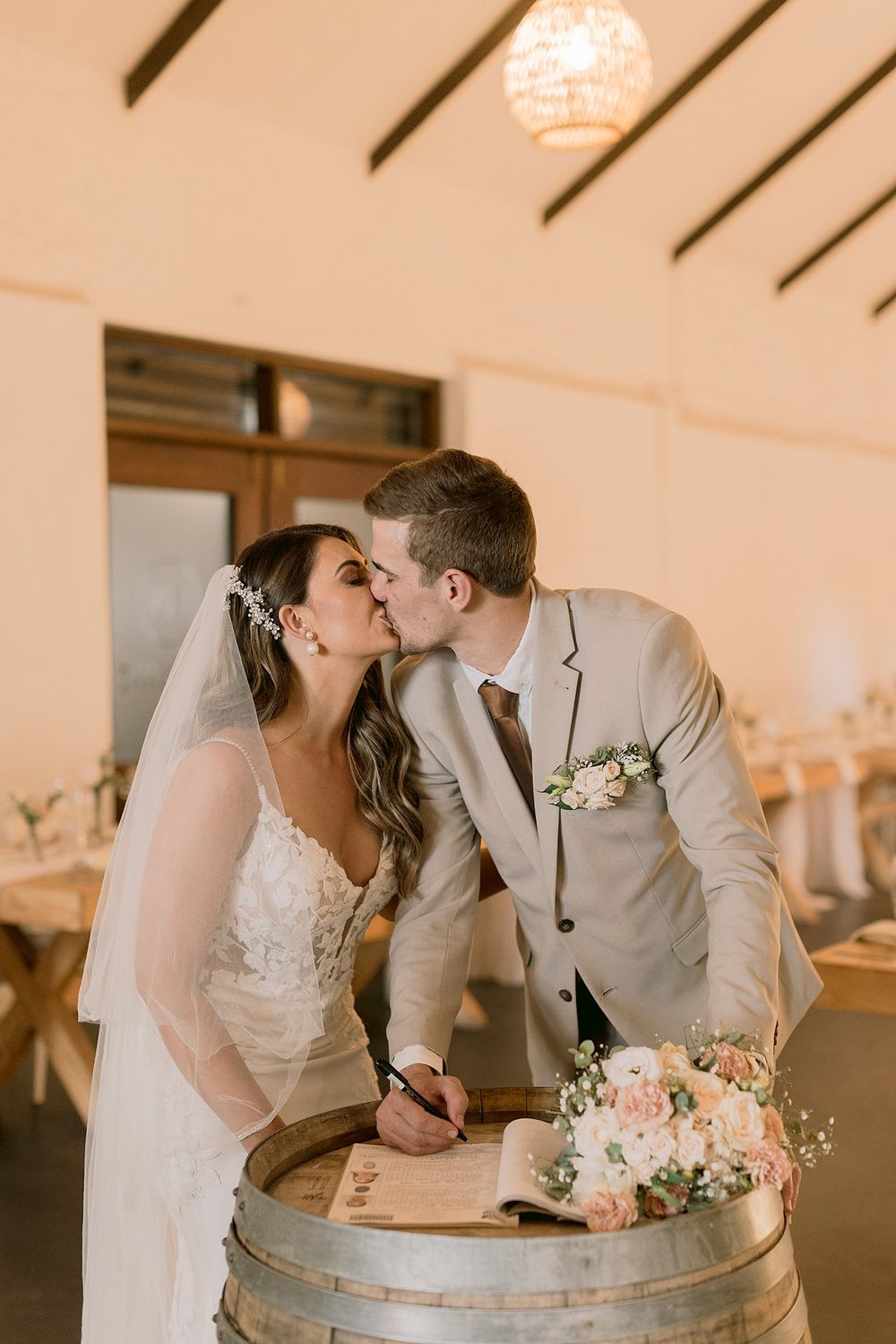 Bride and groom kiss while signing wedding register on a wine barrel in elegant rustic reception venue.
