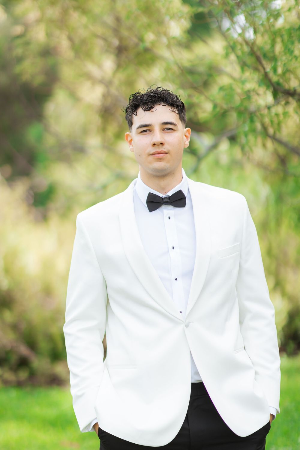 A person in a crisp white dinner jacket and black bow tie poses outdoors against a soft green natural backdrop.