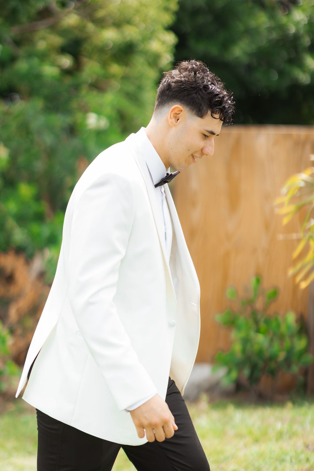 Person in a formal white tuxedo jacket and black bowtie walking outdoors in a garden setting.