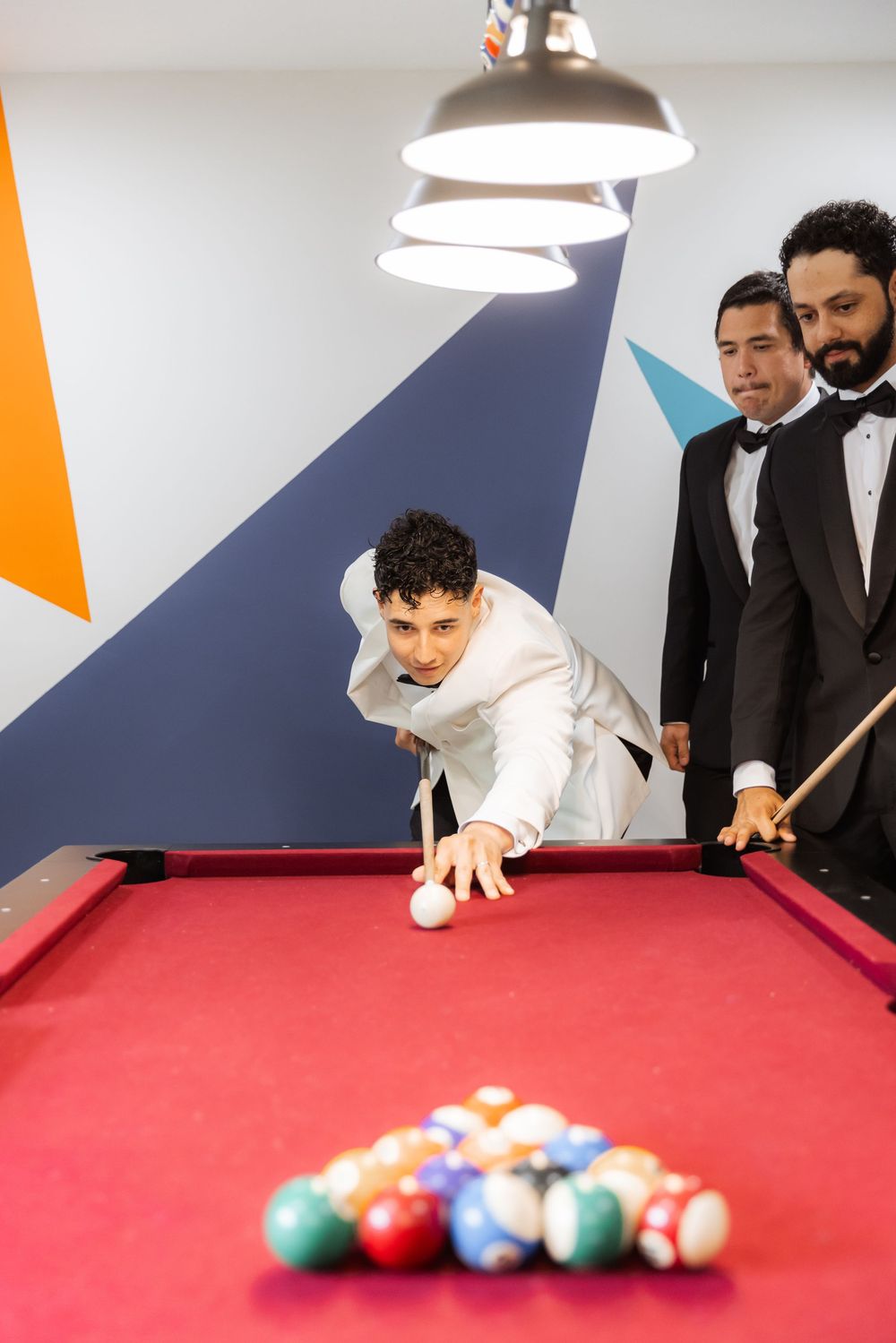Three people in formal attire playing pool on a red table with colorful billiard balls against a geometric wall design.