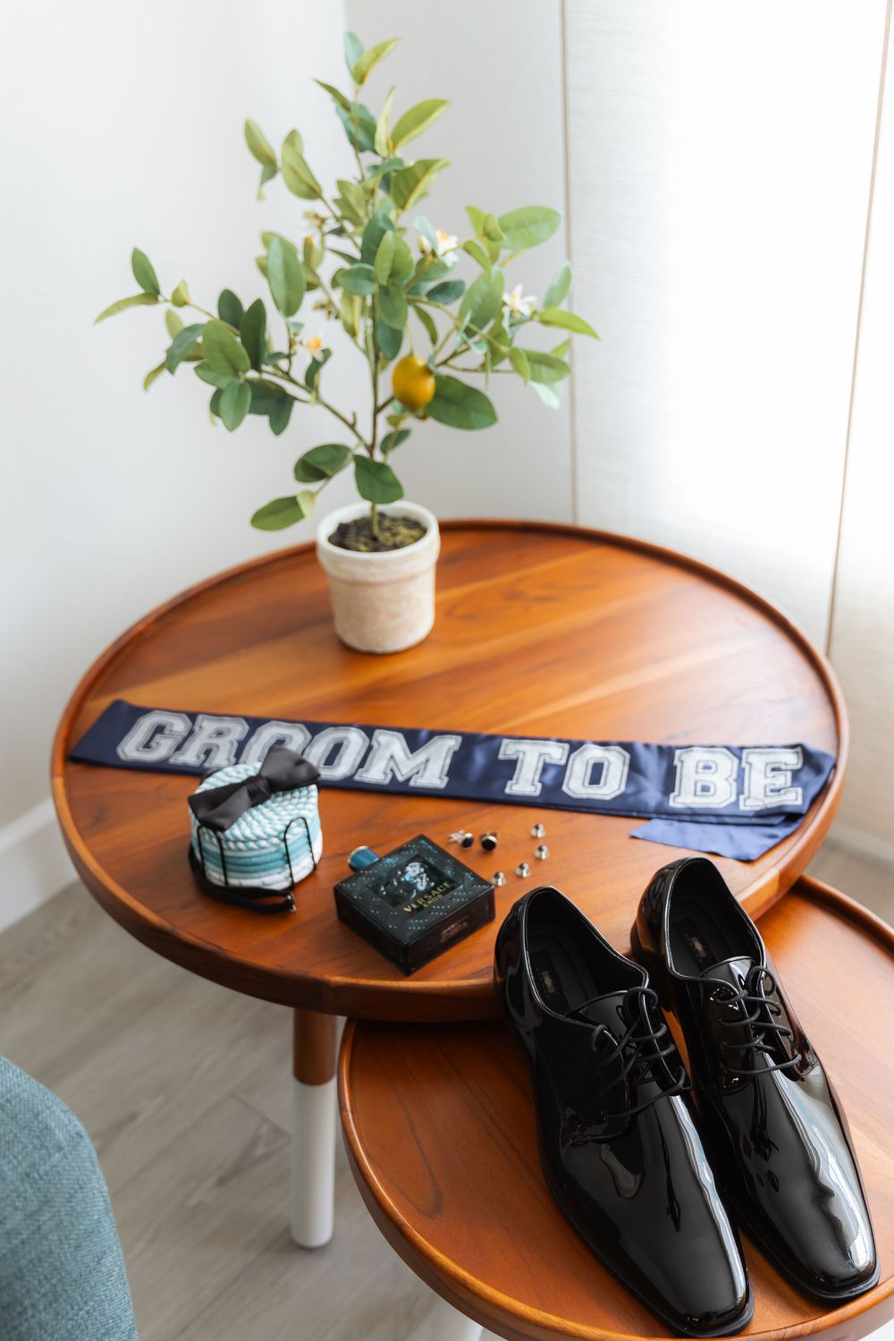 Wedding accessories including groom's patent leather shoes, satin bow tie, and 'Groom to Be' banner on wooden table with small plant.